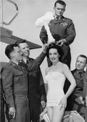 Copa Girl Linda Lawson as "Miss-Cue" wearing an A-Bomb crown to illustrate another misfiring of the Operation Cue Bomb on May 1, 1955. She is surrounded by servicemen. Photo Credit: University of Nevada, Las Vegas Libraries