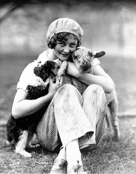 Girl wearing hat and pants seated in the grass holding a lamb and puppy, possibly Washington, ca. 1929-1932 (University of Washington Libraries/History By Zim) Photo Credit: